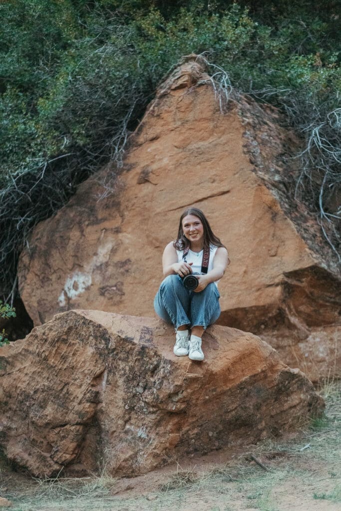 a girl in jeans and white tank top sits on a large red rock holding a camera with another large rock and vines are behind her