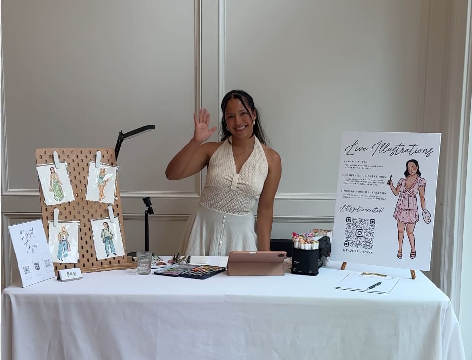 Jasmin Faz, a latina girl stands behind her booth, smiling at the camera, with live watercolor illustrations of people 