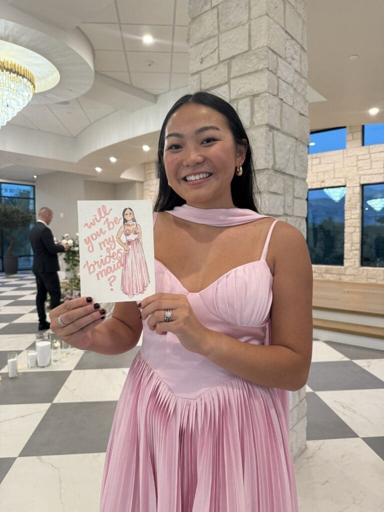 An asian girl in a pink dress holding a water color portrait of herself with the words that say "will you be my bridesmaid?"