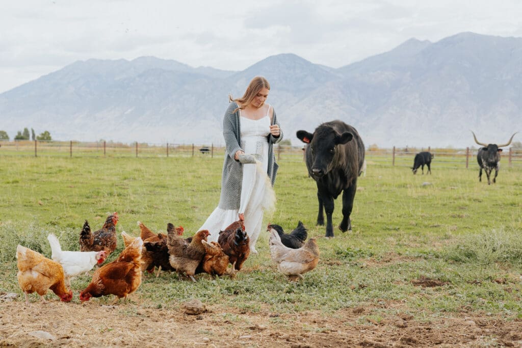 A girl in a flowy dress feeds chickens while a cow approaches her form behind with utah mountains in the background